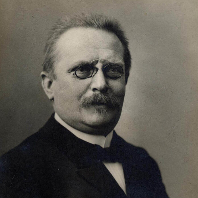 Das Bild zeigt ein historisches Schwarz-Weiß-Porträt von Friedrich Wilhelm Putzger. Dargestellt ist ein Mann mit Schnurrbart und runder Brille, der einen dunklen Anzug mit Fliege trägt.