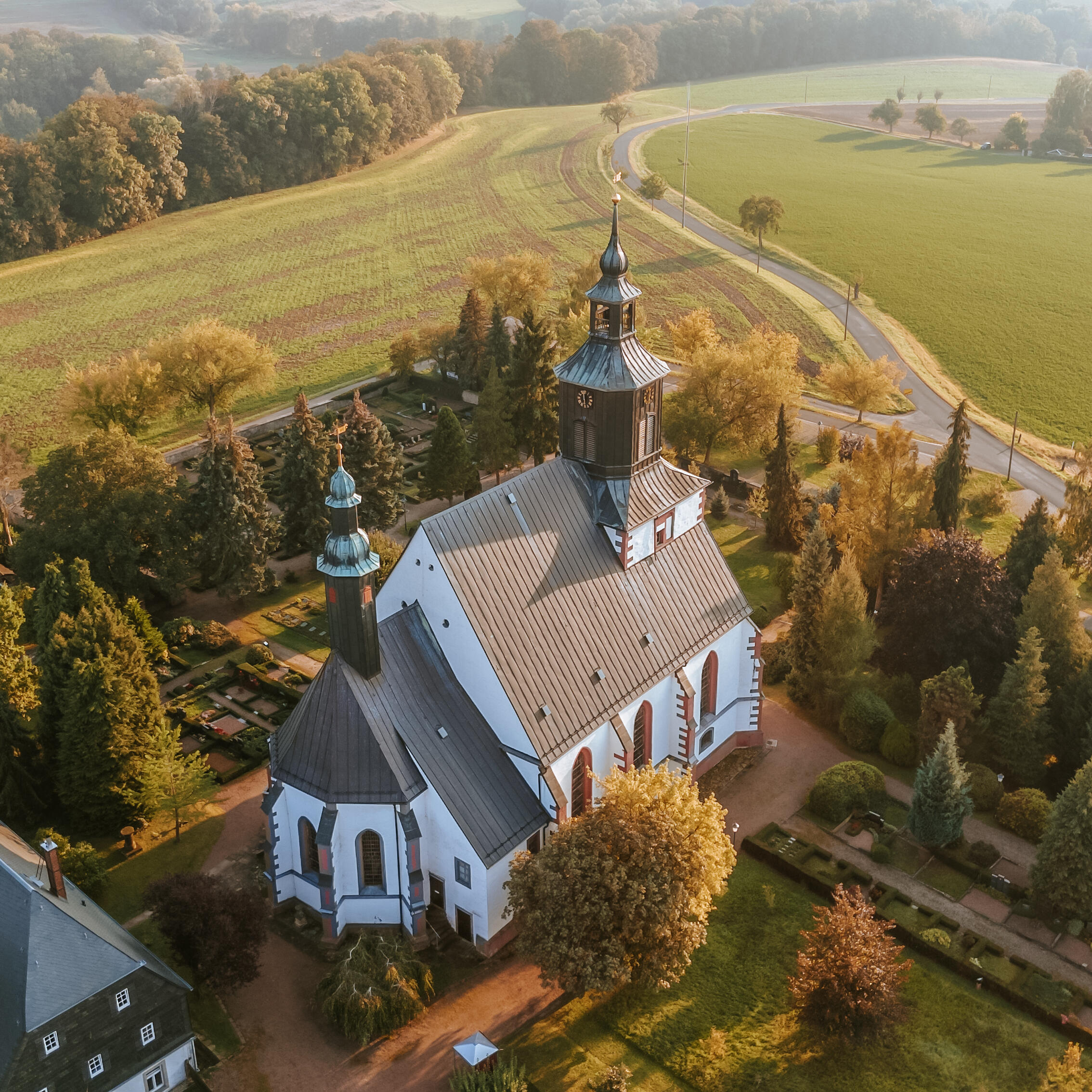 Das Bild zeigt eine Luftaufnahme der Kirche in Seelitz. Die St. Annen Kirche mit zwei Türmen steht inmitten eines Friedhofs mit Bäumen und Grabstätten und ist von Feldern, Wiesen und einer kurvigen Straße in einer ländlichen Landschaft umgeben.