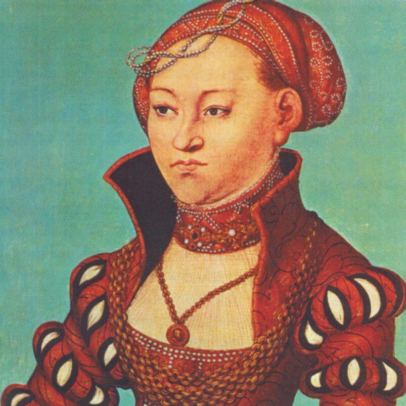 Das Bild zeigt ein historisches Porträt von Elisabeth von Sachsen. Dargestellt ist eine Frau mit roter Kopfbedeckung und reich verzierter roter Kleidung, die Schmuckketten und einen verzierten Kragen trägt und seitlich nach links blickt.