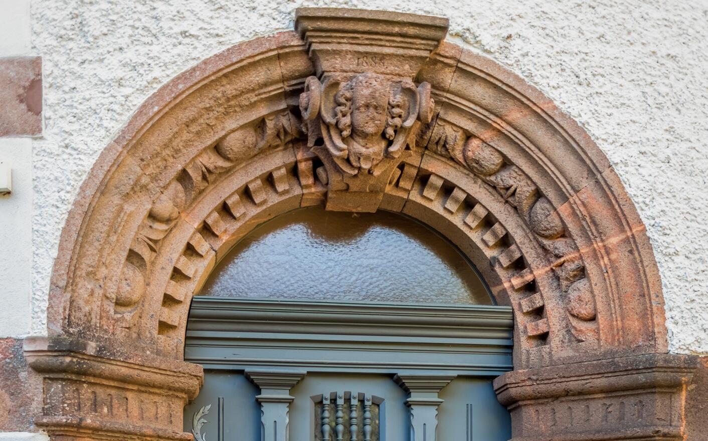 Detailansicht eines Hauseingangs: Eine grau gestrichene Holztür mit kleinem, vergittertem Fenster ist von einem reich verzierten Rundbogen aus rötlichem Porphyrstein umgeben. Im Bogen sind ornamentale Muster und ein Kopfrelief eingearbeitet. Seitlich der Tür befinden sich steinerne Verzierungen in Form von Muscheln sowie kleine, schalenförmige Konsolen.