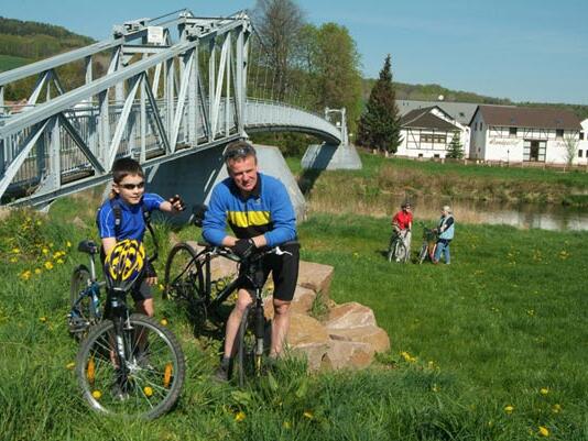 Das Bild zeigt zwei Radfahrer, die ihre Fahrräder auf einer Wiese neben einer Metallbrücke über die Mulde schieben. Im Hintergrund sind weitere Personen mit Fahrrädern sowie Gebäude und Bäume in der Landschaft des Muldentals zu sehen.