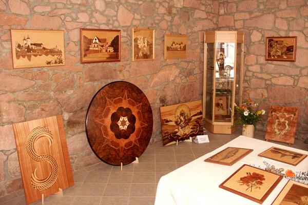 Das Bild zeigt den Ausstellungsraum der Kunstscheune des Vereins „Muldentaler Intarsienkunst e. V.“ im Ortsteil Sörnzig. An den Steinwänden hängen verschiedene Holzkunstwerke, während auf Tischen und am Boden kunstvolle Intarsienarbeiten aus Furnierholz präsentiert werden. In einer Glasvitrine sind weitere Ausstellungsstücke zu sehen.