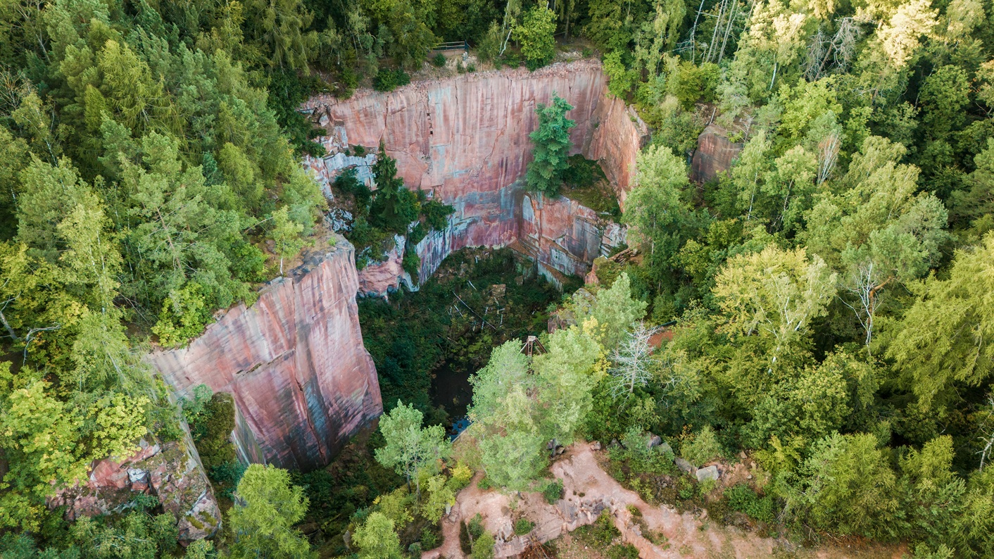 Luftaufnahme des Gleisbergbruchs auf dem Rochlitzer Berg: Ein tiefer, runder Steinbruch mit steilen, rötlichen Felswänden liegt inmitten eines dichten, grünen Waldes. Am Grund des Bruchs befindet sich Wasser und vereinzelte Vegetation. Die umliegenden Bäume reichen bis dicht an den Rand der Felswände heran.
