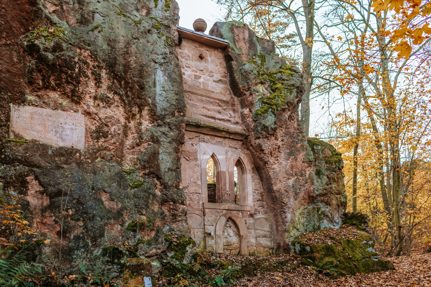 Freistehende historische Steinstruktur aus Rochlitzer Porphyr am Rochlitzer Berg. In den rötlich-grünen Fels ist eine kleine gotische Nischenarchitektur mit Spitzbögen eingelassen. Moos und Herbstlaub bedecken den Boden, im Hintergrund stehen herbstlich gefärbte Laubbäume.