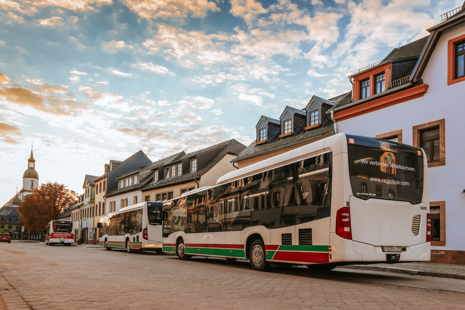 Das Bild zeigt eine Bushaltestelle in der Rathausstraße in Rochlitz. Im Vordergrund stehen mehrere moderne, weiße Reisebusse, die entlang der Straße parken. Die Busse haben grüne und rote Streifen an den Seiten. Im Hintergrund sieht man ein Gebäude mit einer klassischen Architektur, das in hellen Farben gehalten ist. Der Himmel ist weit und klar, mit einigen Wolken. Ein Turm eines kirchlichen Gebäudes ist am linken Rand des Bildes sichtbar. Es ist später Nachmittag oder früher Abend, da das Licht warm und sanft wirkt.