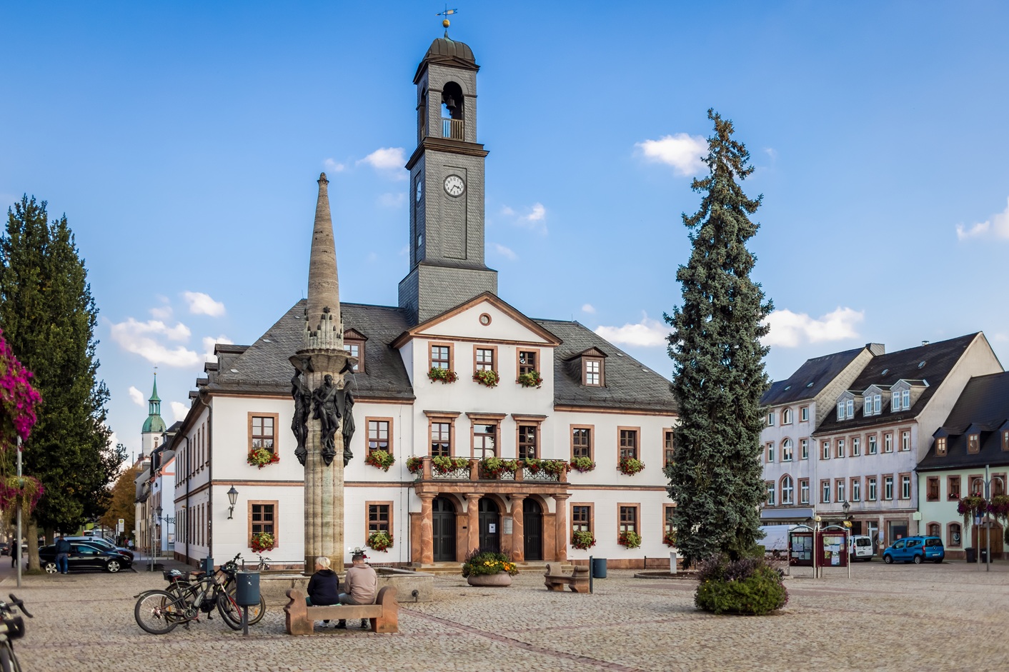 Marktplatz von Rochlitz mit historischem Rathaus: Ein zweigeschossiges Gebäude mit hellem Putz, vielen Fenstern und Blumenkästen steht zentral im Bild. Darüber erhebt sich ein schlanker Uhrturm mit Glockenöffnung. Vor dem Rathaus steht ein hoher Brunnen mit Figuren und einer spitzen Säule. Auf dem gepflasterten Platz sitzen zwei Personen auf einer Bank, daneben stehen Fahrräder. Um den Platz herum befinden sich weitere Wohn- und Geschäftshäuser sowie ein großer Baum.