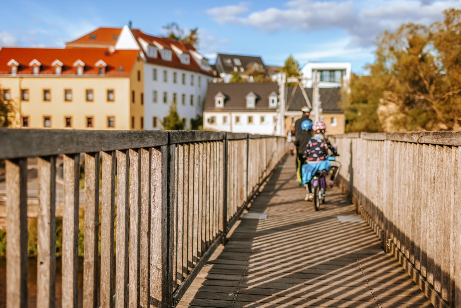 Schaukelbrücke mit Geländer in Rochlitz. Zwei Personen fahren mit Fahrrädern über die Brücke, im Hintergrund stehen Wohnhäuser mit roten und dunklen Dächern. Sonnenlicht wirft lange Schatten auf den Holzbohlen.