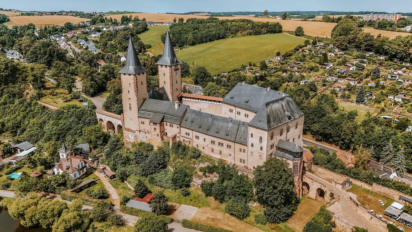 Luftaufnahme von Schloss Rochlitz: Eine große, historische Schlossanlage mit zwei hohen spitz zulaufenden Türmen. Umgeben ist das Schloss von grünen Bäumen, Gärten und Feldern; im Hintergrund sind Wohnhäuser, Ackerflächen und sanfte Hügel der sächsischen Landschaft zu sehen.