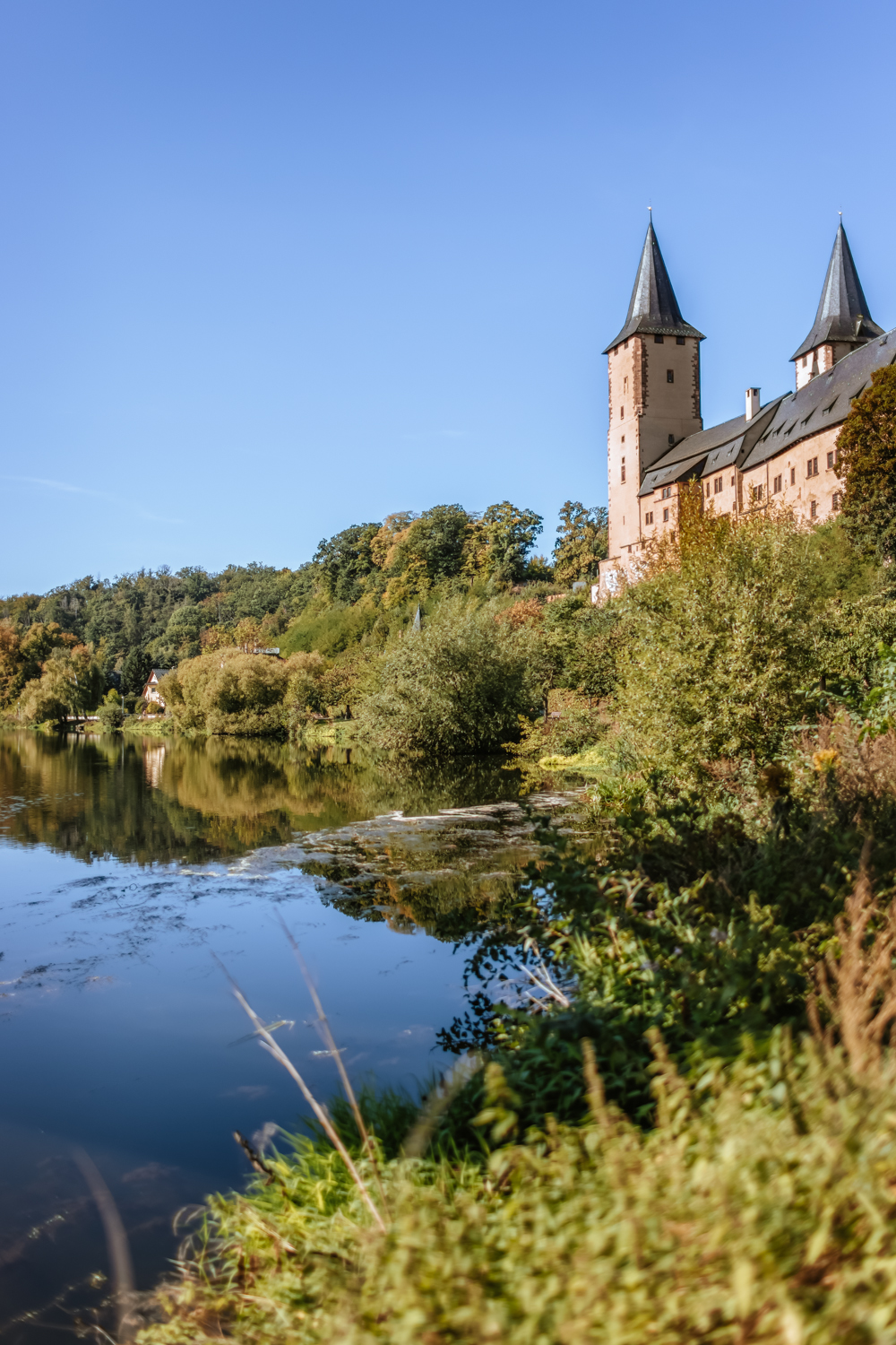 Schloss Rochlitz mit zwei markanten Türmen erhebt sich über der Mulde. Das Schloss spiegelt sich im ruhigen Fluss, umgeben von grünen Bäumen und bewachsener Uferlandschaft bei klarem Himmel.