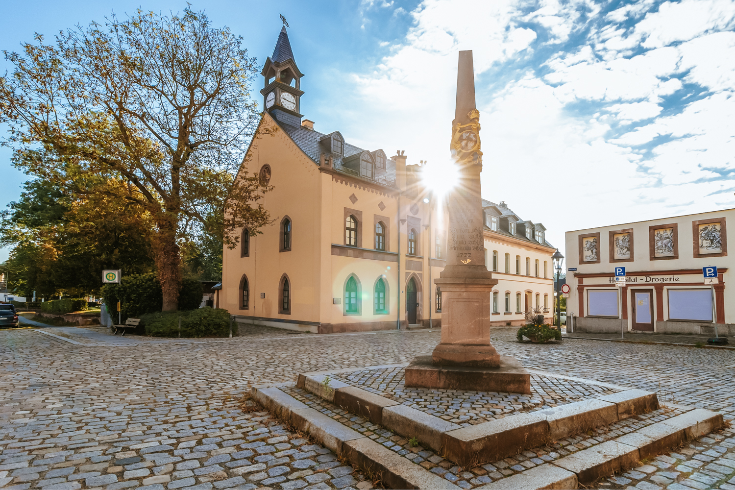 Historischer Marktplatz in Rochlitz (Clemens-Pfau-Platz) mit Kopfsteinpflaster. Im Vordergrund steht ein steinernes Denkmal (Postmeilensäule). Seitlich befinden sich weitere historische Gebäude, die Sonne scheint tief am Himmel.