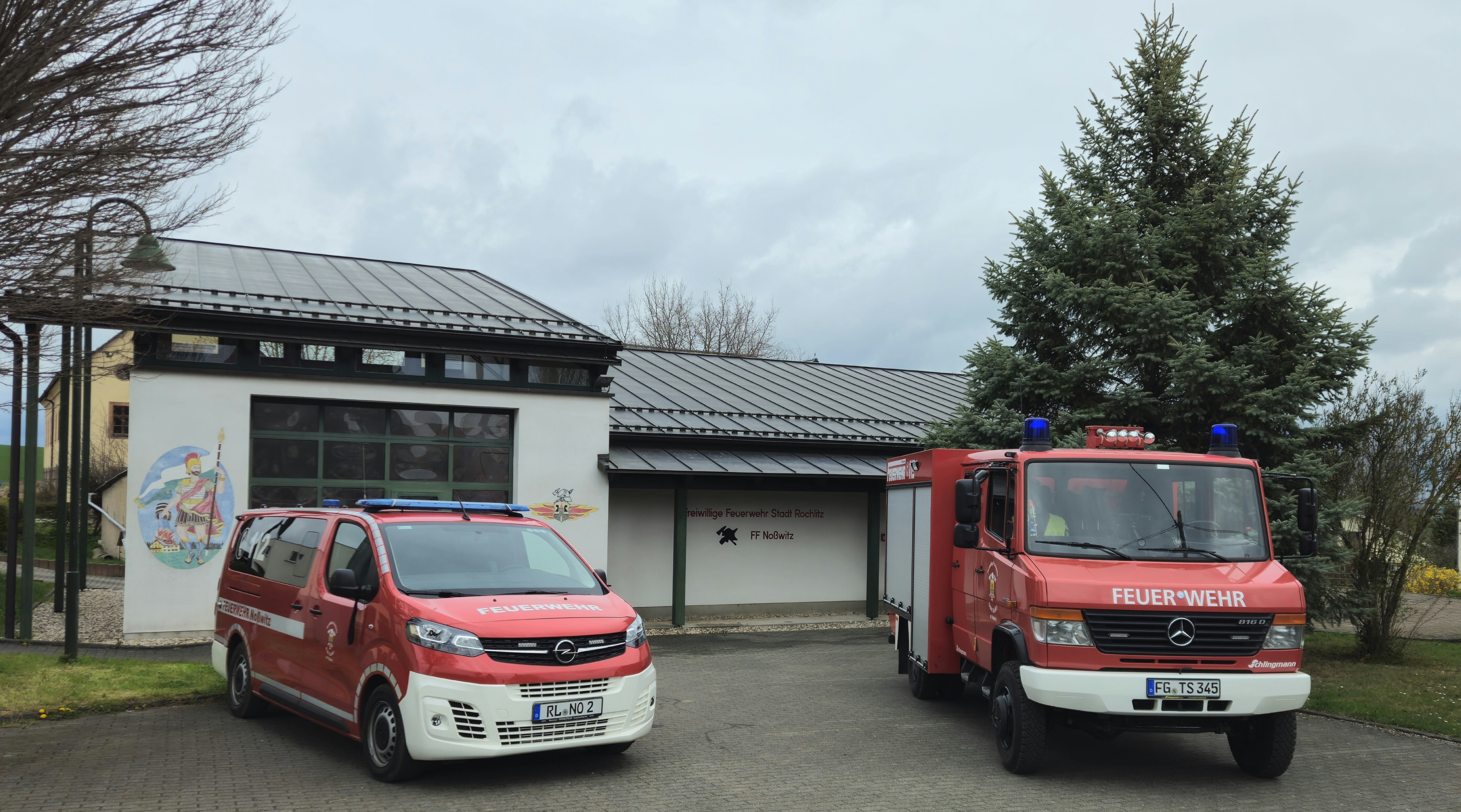 Feuerwehrgerätehaus der Freiwilligen Feuerwehr Noßwitz mit zwei roten Einsatzfahrzeugen vor dem Gebäude. Im Hintergrund stehen Tannen und ein bewölkter Himmel