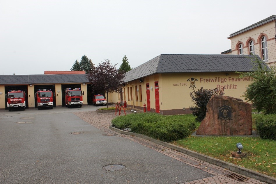 Gelände der Freiwilligen Feuerwehr Rochlitz mit mehreren Einsatzfahrzeugen vor den Fahrzeughallen. Vor dem Gebäude steht ein Gedenkstein mit Feuerwehrwappen.
