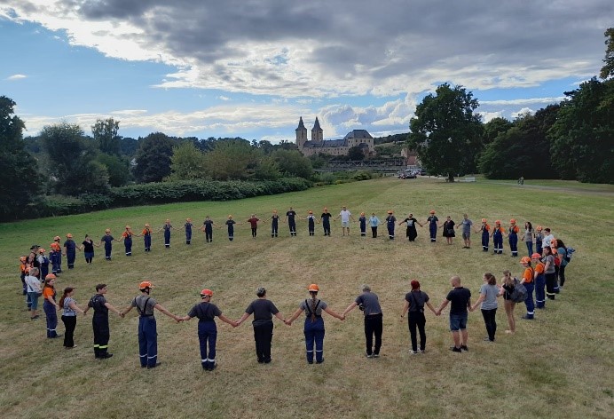 Mitglieder der Jugendfeuerwehr und Betreuende stehen im Kreis auf einer Wiese und halten sich an den Händen. Im Hintergrund ist Schloss Rochlitz zu sehen