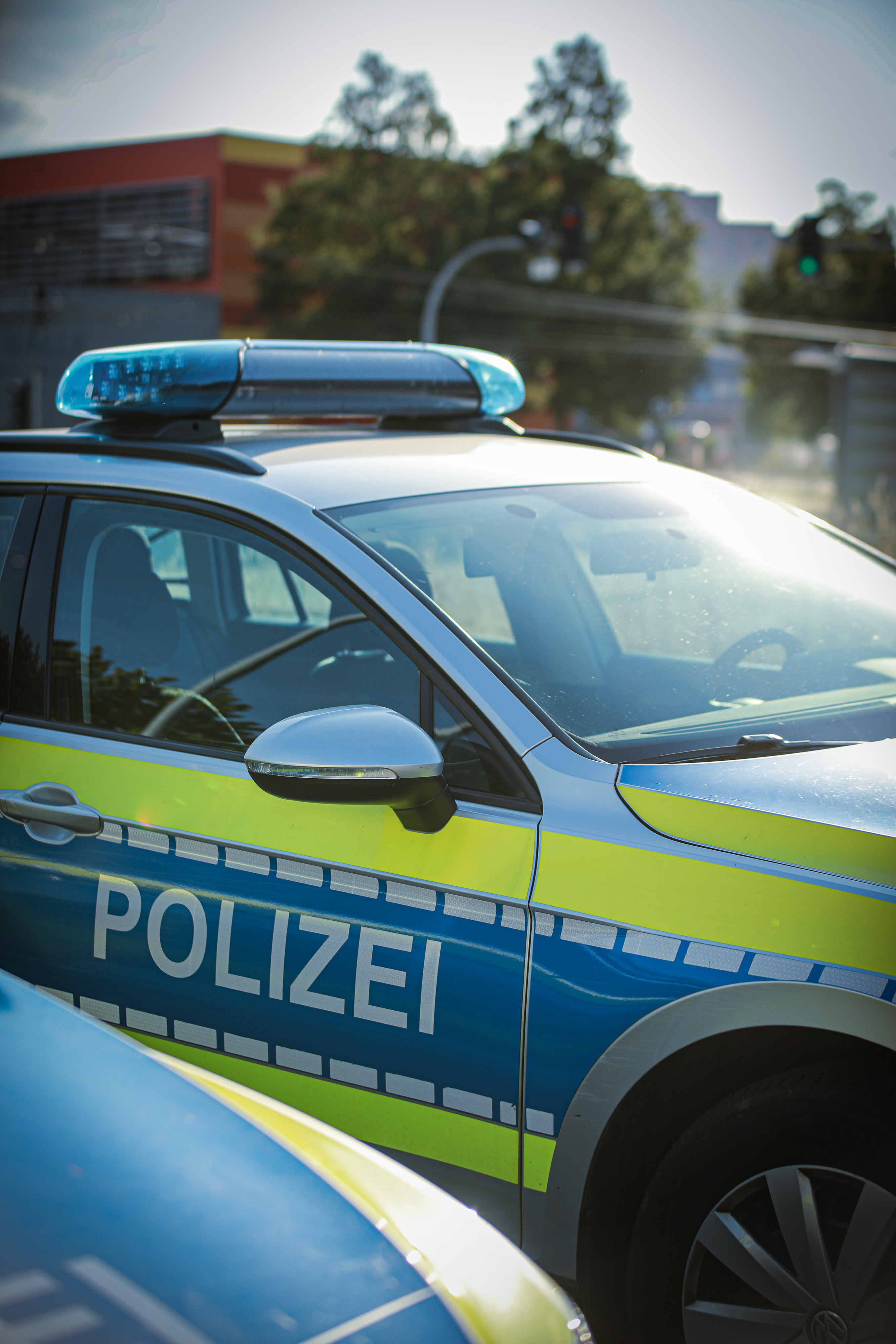 Ein Polizeifahrzeug mit blauem Einsatzlicht auf dem Dach steht im Straßenverkehr. Auf der Fahrzeugseite ist der Schriftzug „Polizei“ zu sehen, im Hintergrund befinden sich Gebäude und eine Ampelkreuzung.